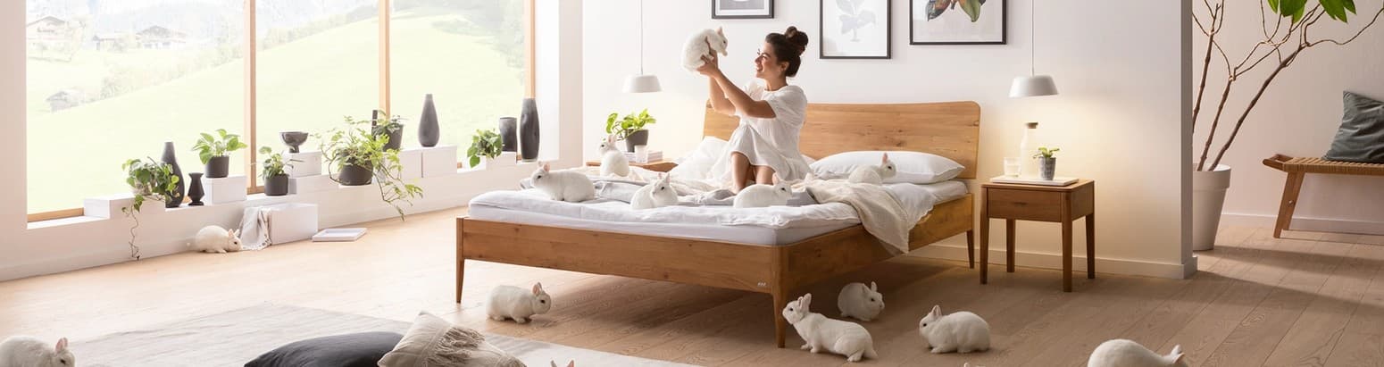 Eine Frau sitzt auf einem Massivholzbett im Schlafzimmer und hält ein Kaninchen in die Luft. Weitere Kaninchen umgeben sie auf dem Bett und im Raum.